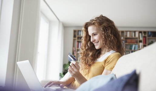 Junge Frau mit lockigem, rotbraunem Haar sitzt lächelnd auf dem Sofa und hält eine Kreditkarte vor dem Laptop in einem hellen Wohnzimmer. | © Getty Images/Westend61