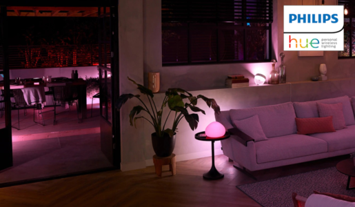 Wohnzimmer mit violettem Ambientelicht, grauem Sofa, Beistelltisch mit rosa Lampe und Blick auf beleuchtete Terrasse bei Nacht. | © Amazon/Philips