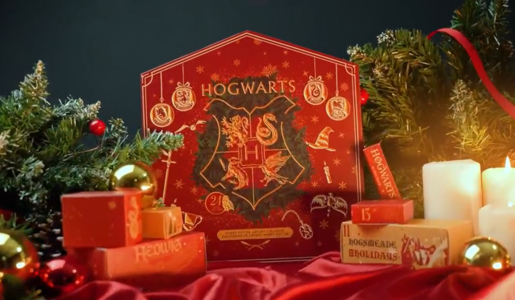 Roter Harry Potter-Adventskalender mit Hogwarts-Wappen, umgeben von Tannenzweigen, Kerzen und goldenen Weihnachtskugeln auf rotem Stoff. | © Amazon/Cinereplicas