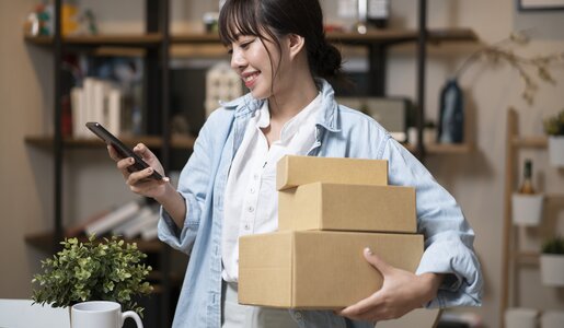 Eine junge Frau mit braunem Zopf und Jeanshemd lächelt, hält drei Kartons und tippt in einem hellen Arbeitszimmer auf ihr Smartphone. | © Getty Images/seksan Mongkhonkhamsao
