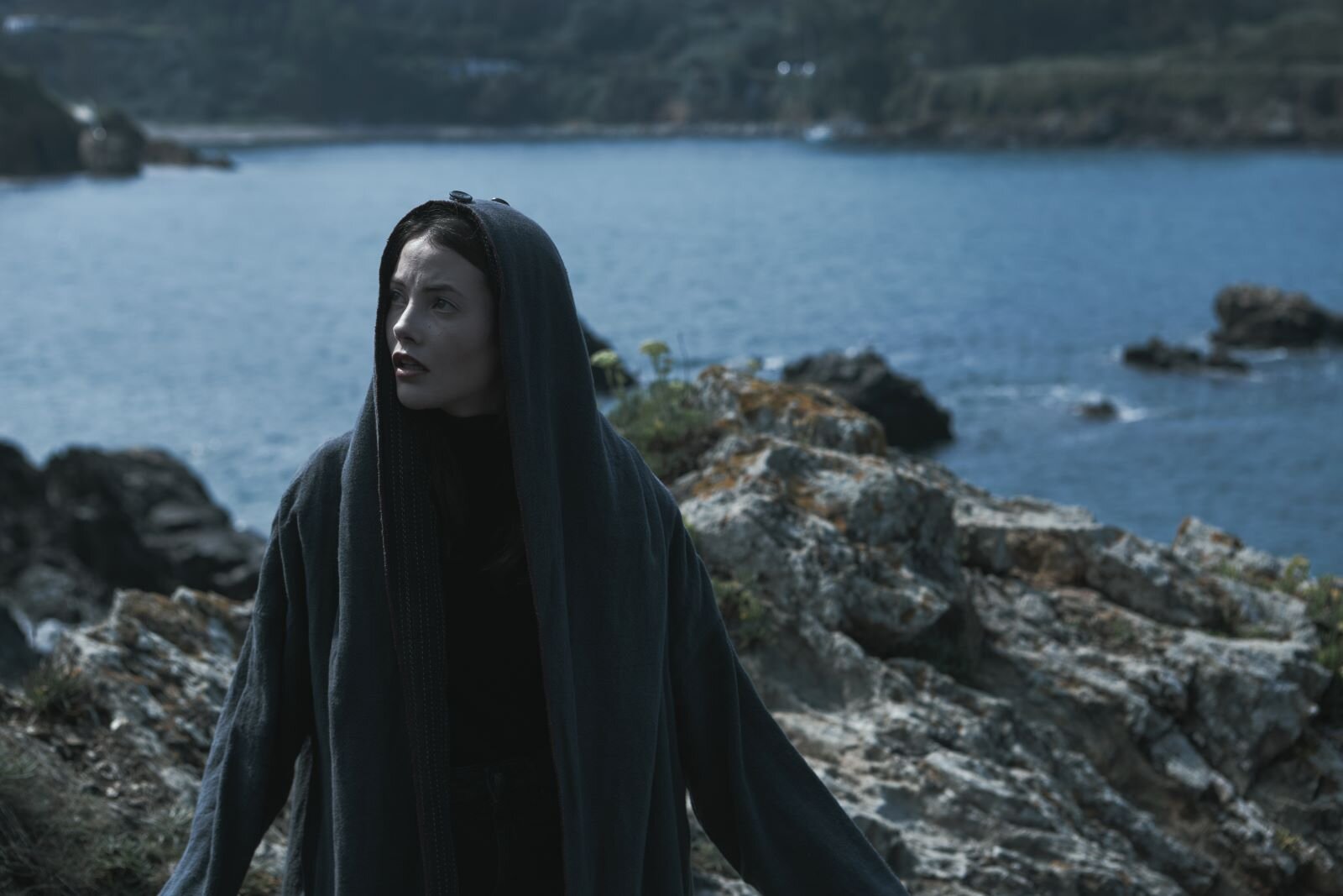MIREIA ORIOL as ALMA in episode 07 of ALMA: Junge Frau mit heller Haut und dunklen Haaren im langen, grauen Umhang blickt besorgt auf felsiger Küste mit blauem Meer. | © ALMA LARRAÑAGA/NETFLIX