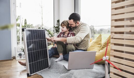 Vater und Sohn betrachten lernend ein Solarmodul neben einem Laptop im Wohnzimmer. | © Getty Images/Westend61