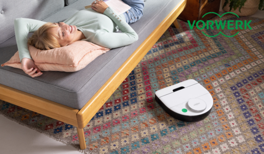Lächelnde junge Frau liegt auf Couch, der Vorwerk Kobold RK7 Saugroboter arbeitet auf gemustertem Teppich im Wohnzimmer. | © Vorwerk