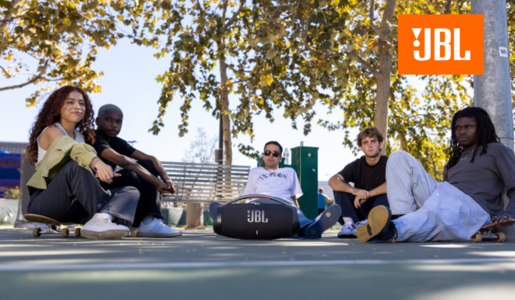 Junge Menschen sitzen rund um die JBL Boombox, dahinter ein Baum und eine Parkbank. | © JBL