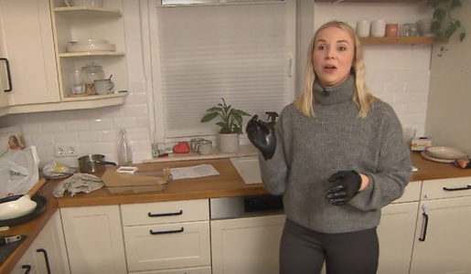 Eine junge Frau mit blonden Haaren steht in einer modernen Küche, trägt einen grauen Pullover und schwarze Handschuhe. | © Screenshot Vox/RTL+ 