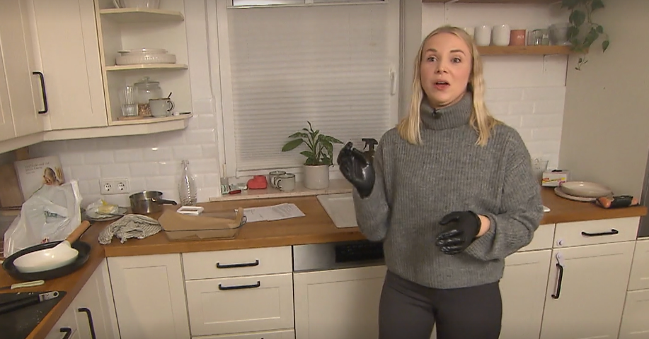 Eine junge Frau mit blonden Haaren steht in einer modernen Küche, trägt einen grauen Pullover und schwarze Handschuhe. | © Screenshot Vox/RTL+ 