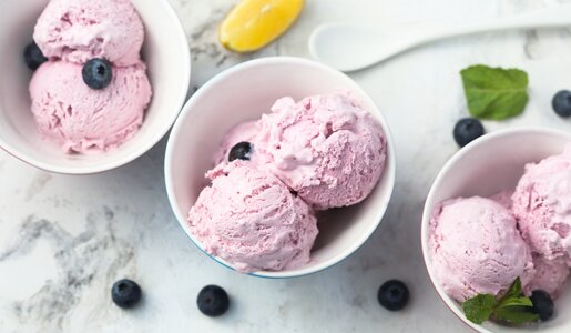 Drei Schalen mit rosa Heidelbeer-Eis, garniert mit Blaubeeren und Minzblättern, auf weißem Marmortisch. | © Getty Images/Mariha-kitchen
