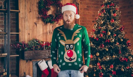 Ein bärtiger Mann in Rentierpullover und Weihnachtsmütze steht vor einem festlich dekorierten Raum mit Weihnachtsbaum und Kamin. | © deagreez/stock.adobe.com 