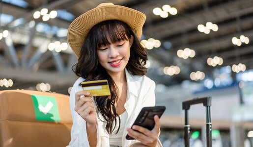 Junge Frau mit Strohhut und langen dunklen Haaren ist am Flughafen und hält lächelnd eine Kreditkarte und ein Smartphone in der Hand. | © Getty Images/Twenty47studio