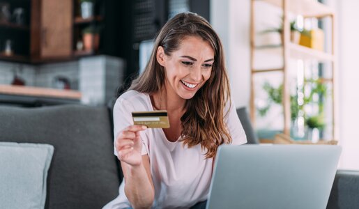 Eine lachende Frau mit braunen Haaren hält eine Kreditkarte in der Hand und sitzt vor einem Laptop im Wohnzimmer. | © Getty Images/VioletaStoimenova